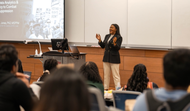 Dr. Chelsea Jones stands in front of an audience, presenting information about voting rights, with her hands raised in front of her as she speaks.